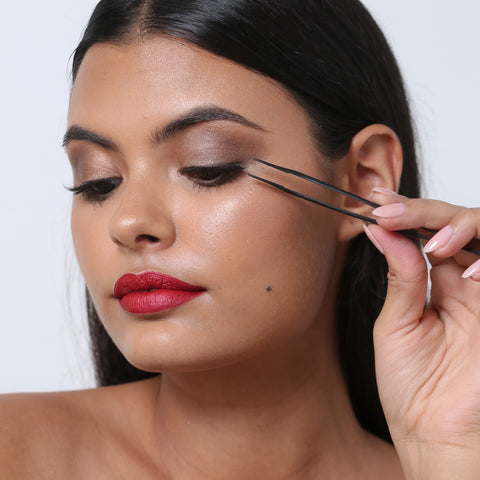 Lithe Lashes false lashes accessories, Lash Applicator. A matte black and ergonomic applicator tool to help in the false lashes application process. The image shows a model holding the applicator in her left hand positioning it by her left eye as she finishes the touch ups from her lash application.