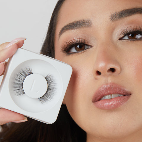 Lithe Lashes Core Collection lash style named 04 Long and Graceful, an image of a model in full makeup, wearing lithe false lashes in both eyes, displaying how elegant they look, while holding up near her right eye and right cheek another pair of the same false lashes still in their biodegradable plastic tray, showcasing the beauty of how the lashes look in their packaging.