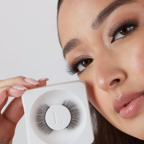 Lithe Lashes Core Collection lash style named 07 Long and 3-Dimensional, an image of a model in full makeup, wearing lithe false lashes in both eyes, displaying how elegant they look, while holding up near her right eye and right cheek another pair of the same false lashes still in their biodegradable plastic tray, showcasing the beauty of how the lashes look in their packaging.