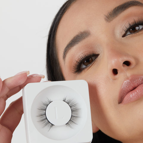 Lithe Lashes Core Collection lash style named 05 Wispy and Willowy, an image of a model in full makeup, wearing lithe false lashes in both eyes, displaying how elegant they look, while holding up near her right eye and right cheek another pair of the same false lashes still in their biodegradable plastic tray, showcasing the beauty of how the lashes look in their packaging.