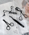 Lithe Lashes false lashes accessory, the Lithe Lash Scissors. A product image showing all Lithe's matte black lash tools, with the Lash Scissors in the bottom left, the Mini Lash Curler at the top, the Lash Applicator at the bottom of the page, and the Lithe Lash Curler in the upper right portion of the image, all of which are laying gently on black and white magazine print.