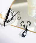 Lithe Lashes false lashes accessory, the Lithe Lash Curler. A product image showing the matte black Lash Applicator and the Mini Lash Curler resting on a small white porcelain plate, while the ergonomic Lithe Lash Curler is resting half on a mirror with thin golden frame and the other half on a spotless white table. In the background is an expensive bottle of perfume and crystal candle holder.
